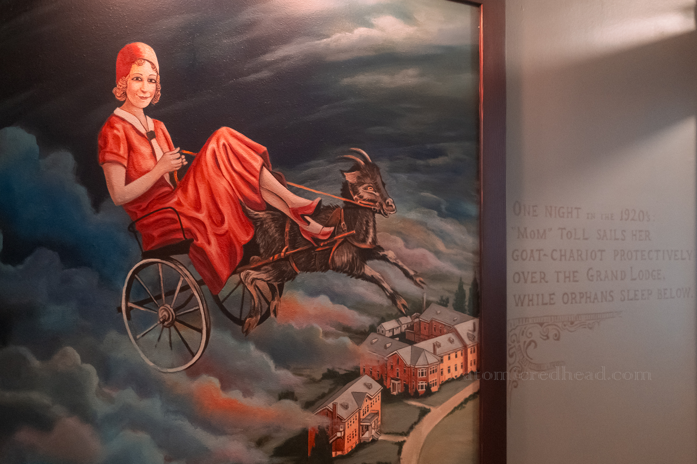 A mural of a woman in a pink 1920s dress sits in a chariot pulled by a goat through the sky above the hotel, to the side it reads "One night in the 1920s: 'Mom' Toll sails her goat-chariot protectively over the Grand Lodge while orphans sleep below."