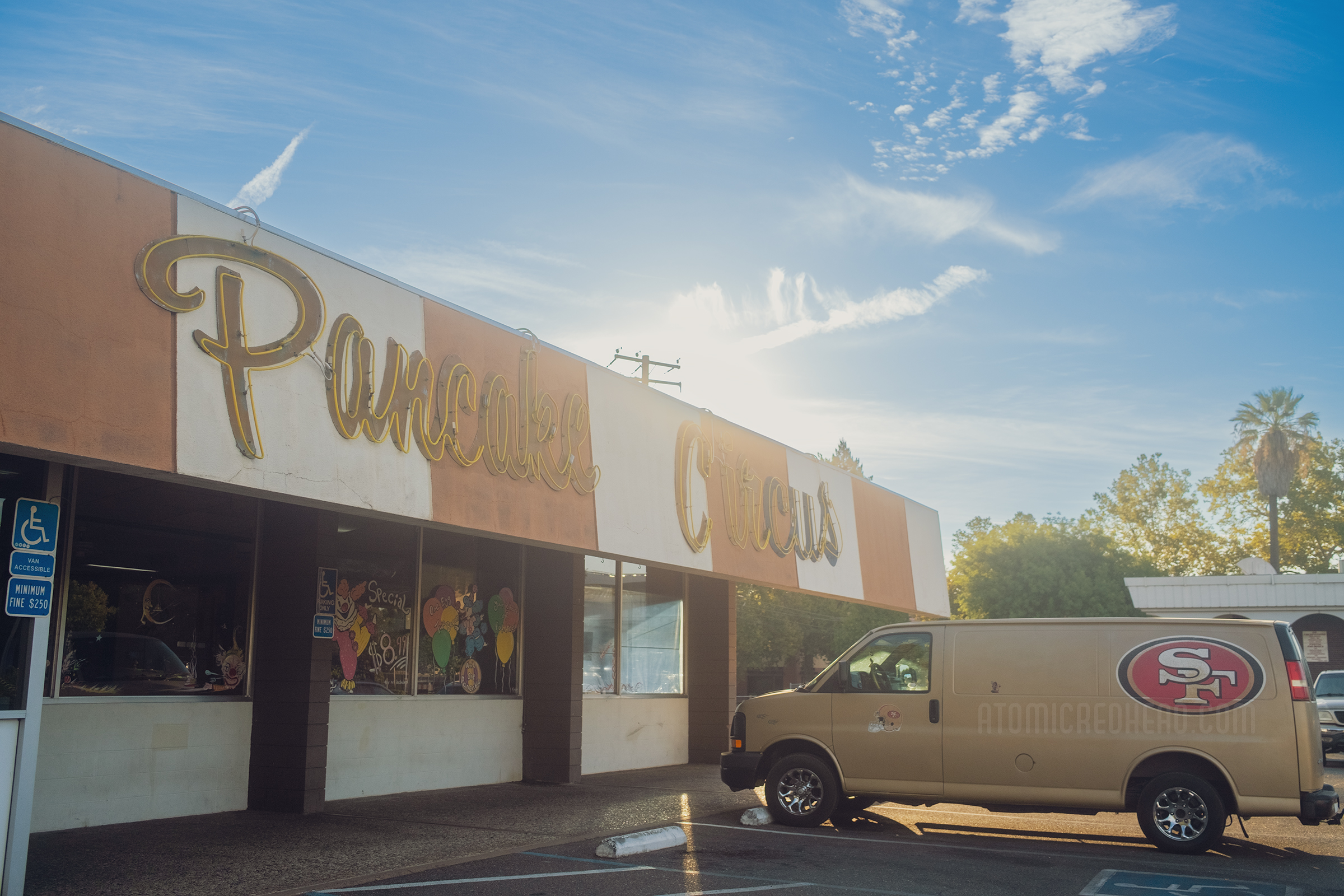 Exterior of Pancake Circus. Across the top the roof comes down and painted in orange and white stripes, yellow script neon reads "Pancake Circus" below massive windows are painted in circus themes.