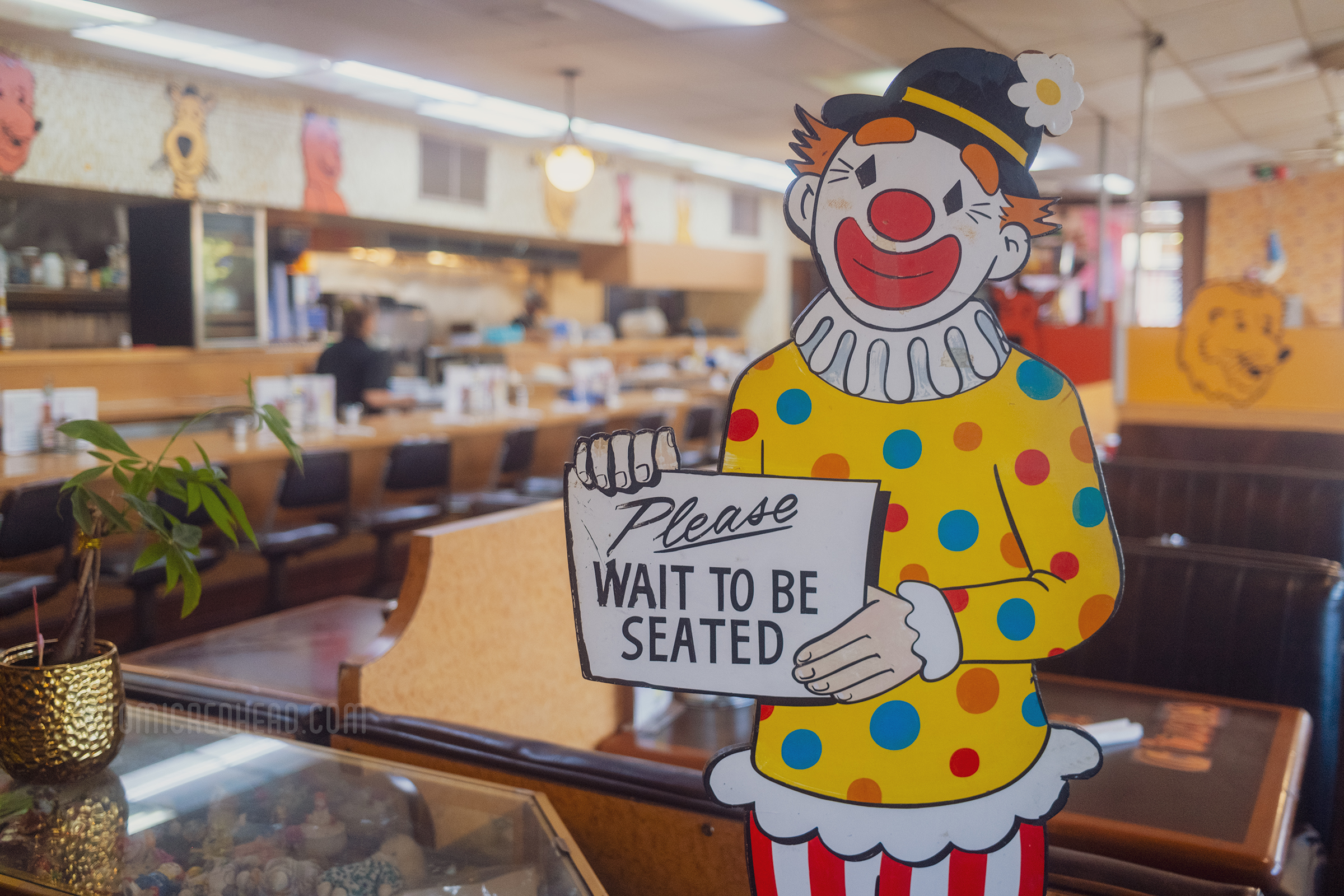 A painted cut out of a clown reads "Please wait to be seated"