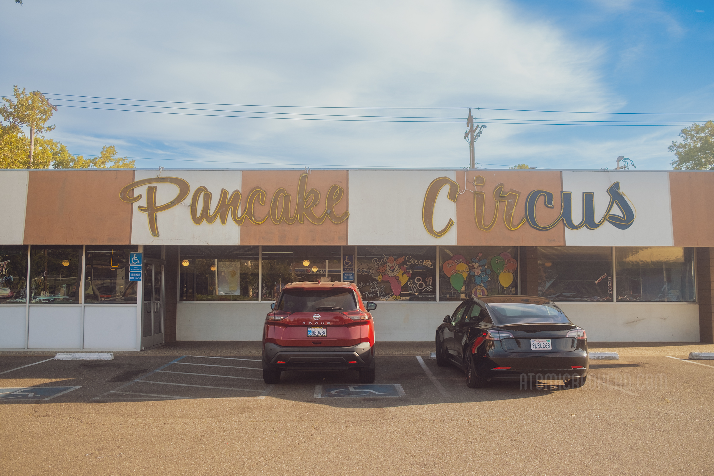Exterior of Pancake Circus. Across the top the roof comes down and painted in orange and white stripes, yellow script neon reads "Pancake Circus" below massive windows are painted in circus themes.