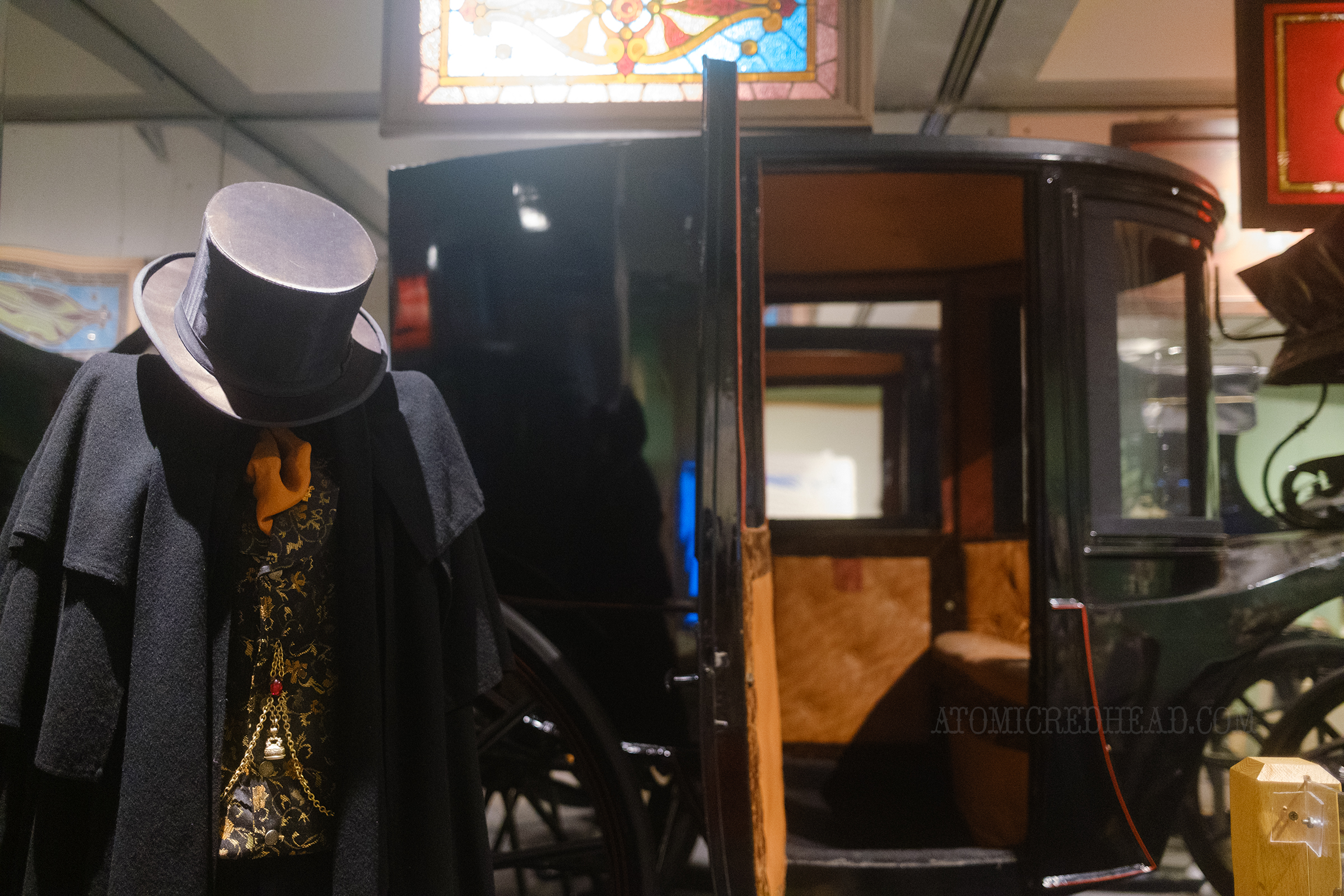 A mannequin features Victorian attire of a well off man ready for an evening out, with a cloak and top hat, behind is a black carriage.
