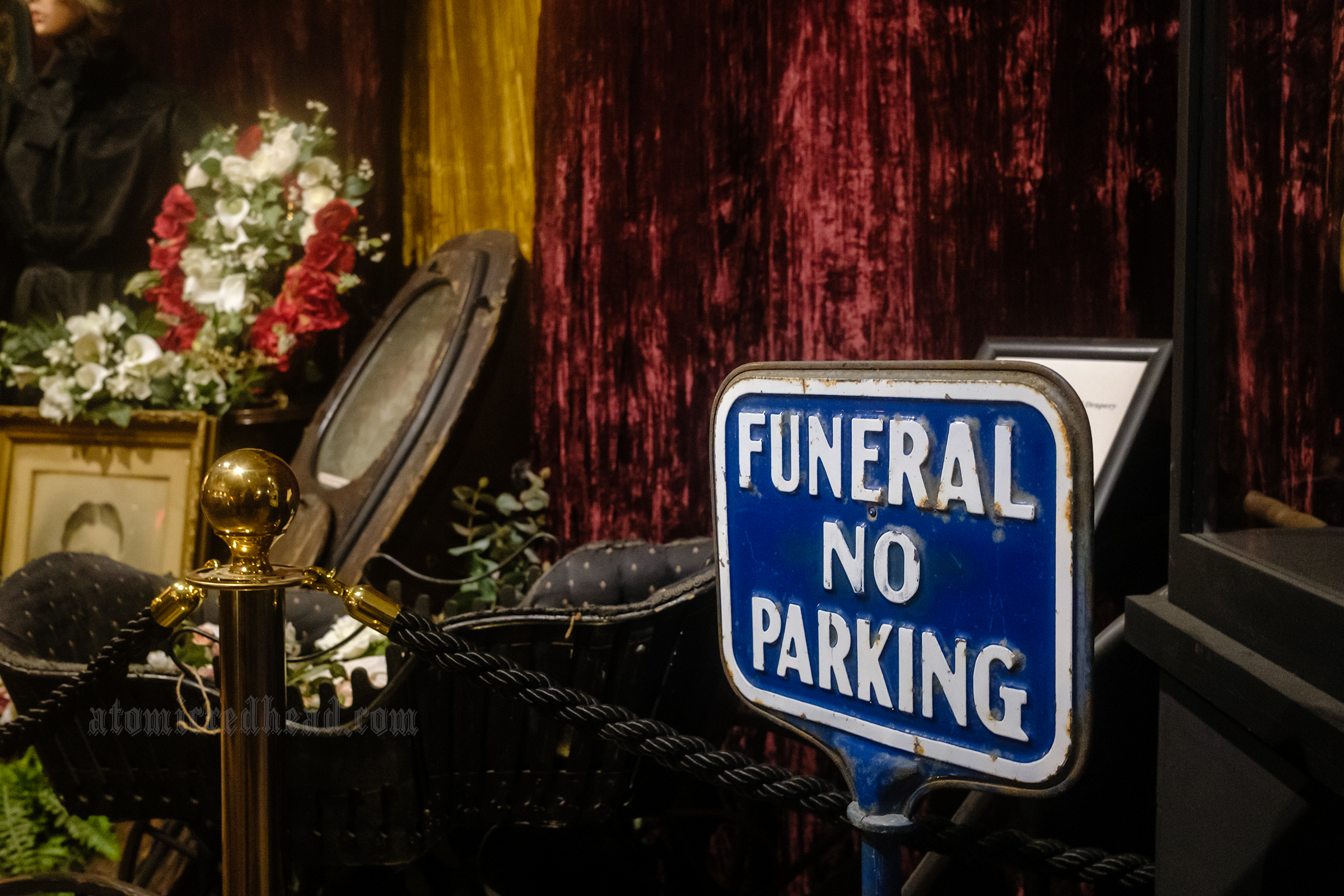 Various coffins rest against a red velvet curtain, on the right a blue and white sign reads "Funeral No Parking"
