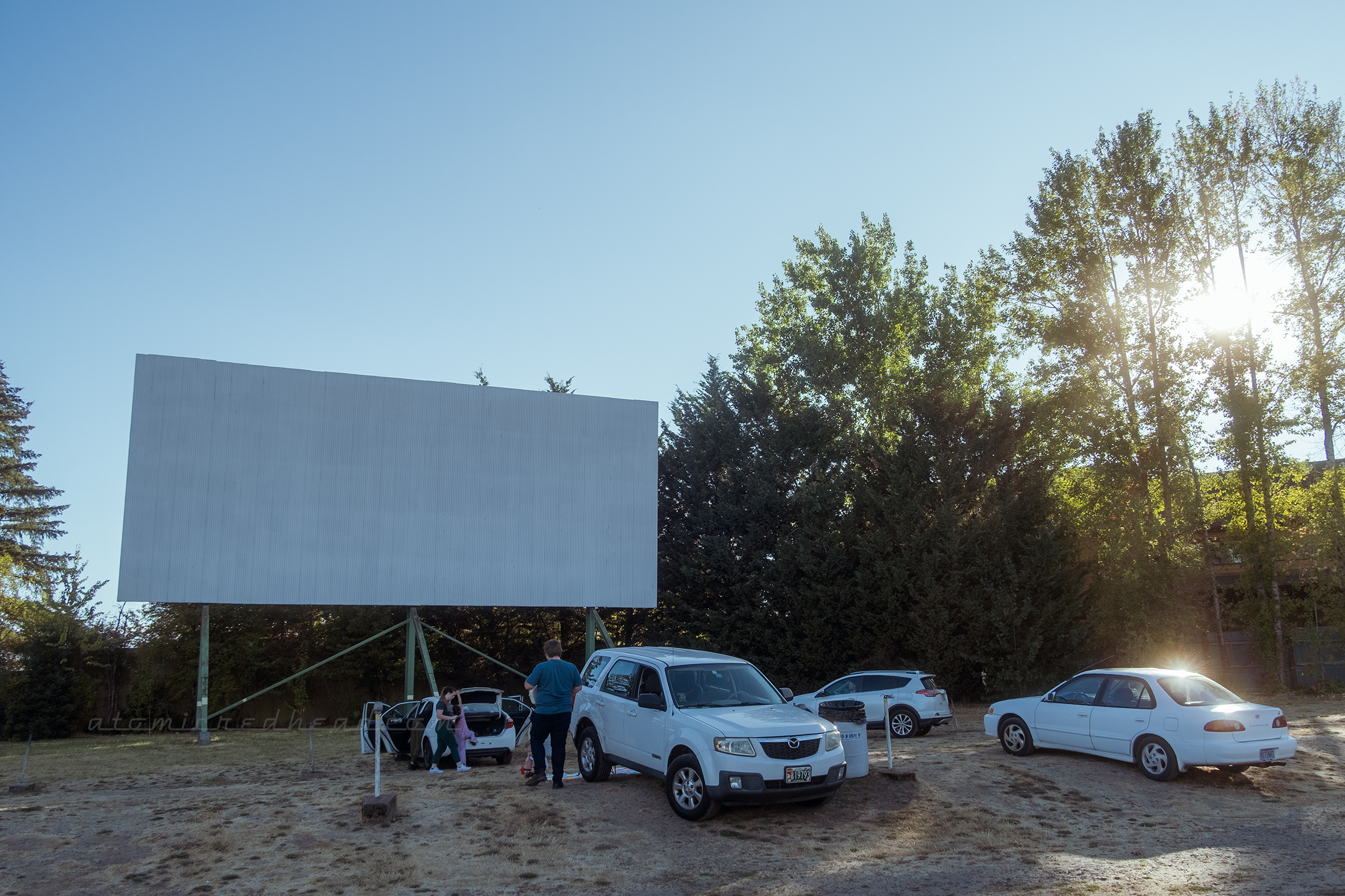 Cars line up in front of the large outdoor screen, with the sun beginning to set behind the trees to the right.