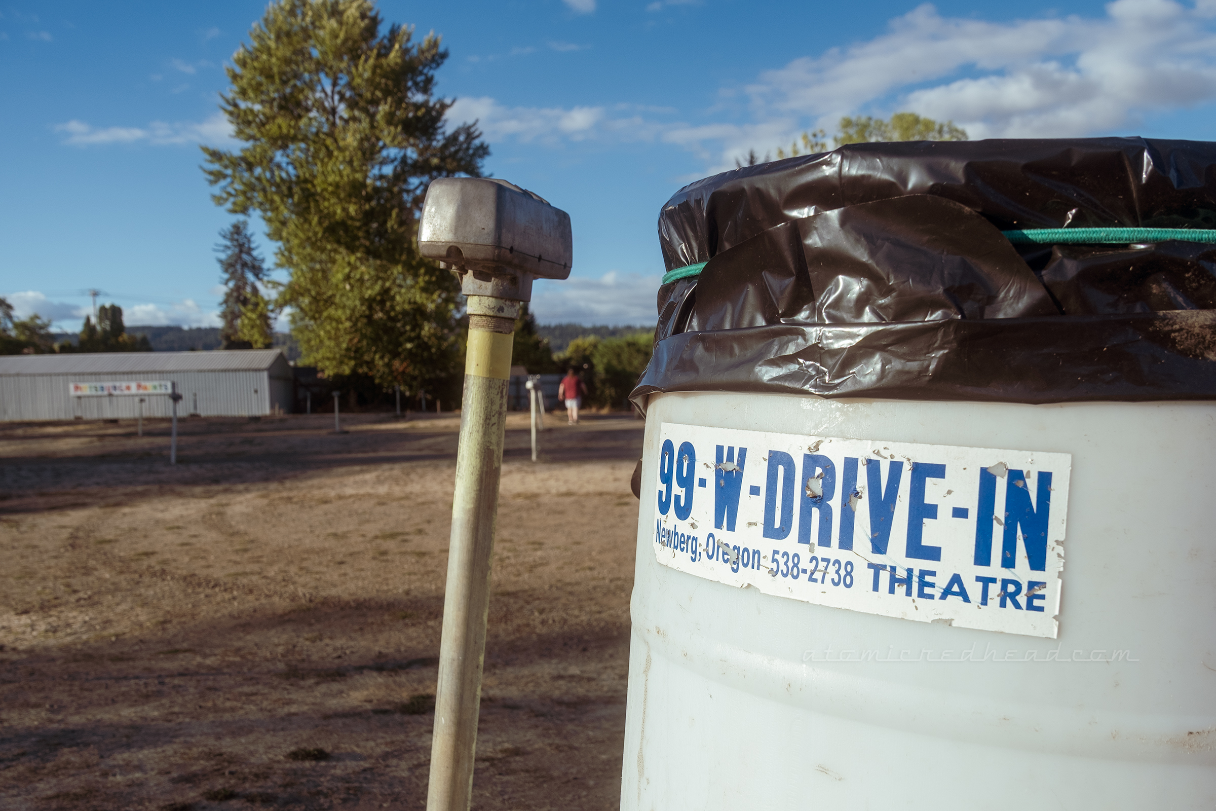 A trash can has a 99W bumper sticker on it, with an old speaker stand next to it.