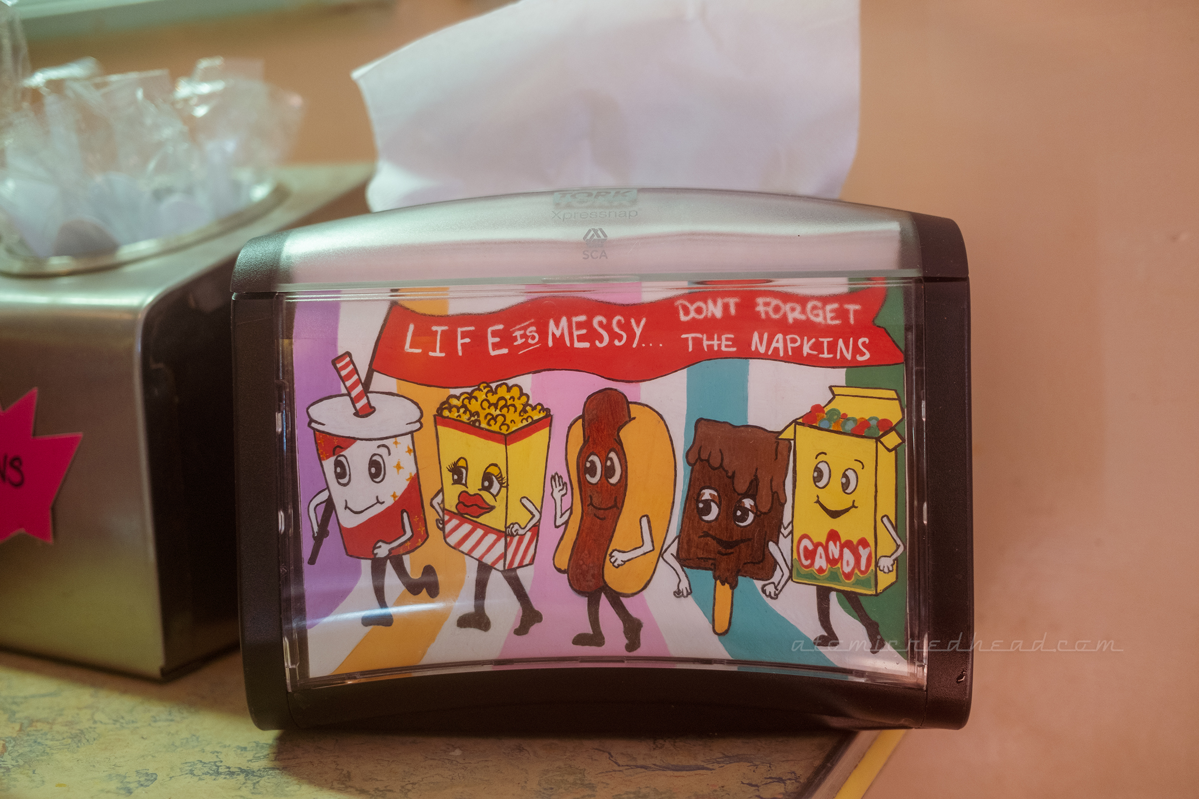 A napkin holder decorated with anthropomorphic candy and popcorn and a banner reading "Life is Messy...don't forget the napkins"