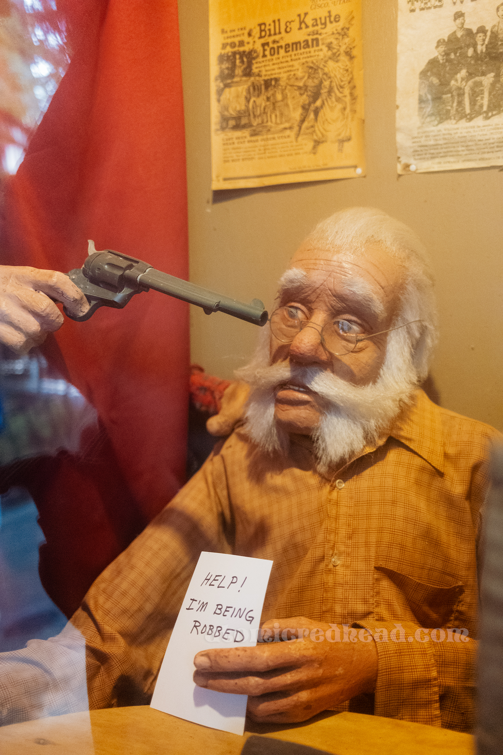 An old man seen through a window in the western town has a gun to his head and he holds a note reading "Help! I'm being robbed"