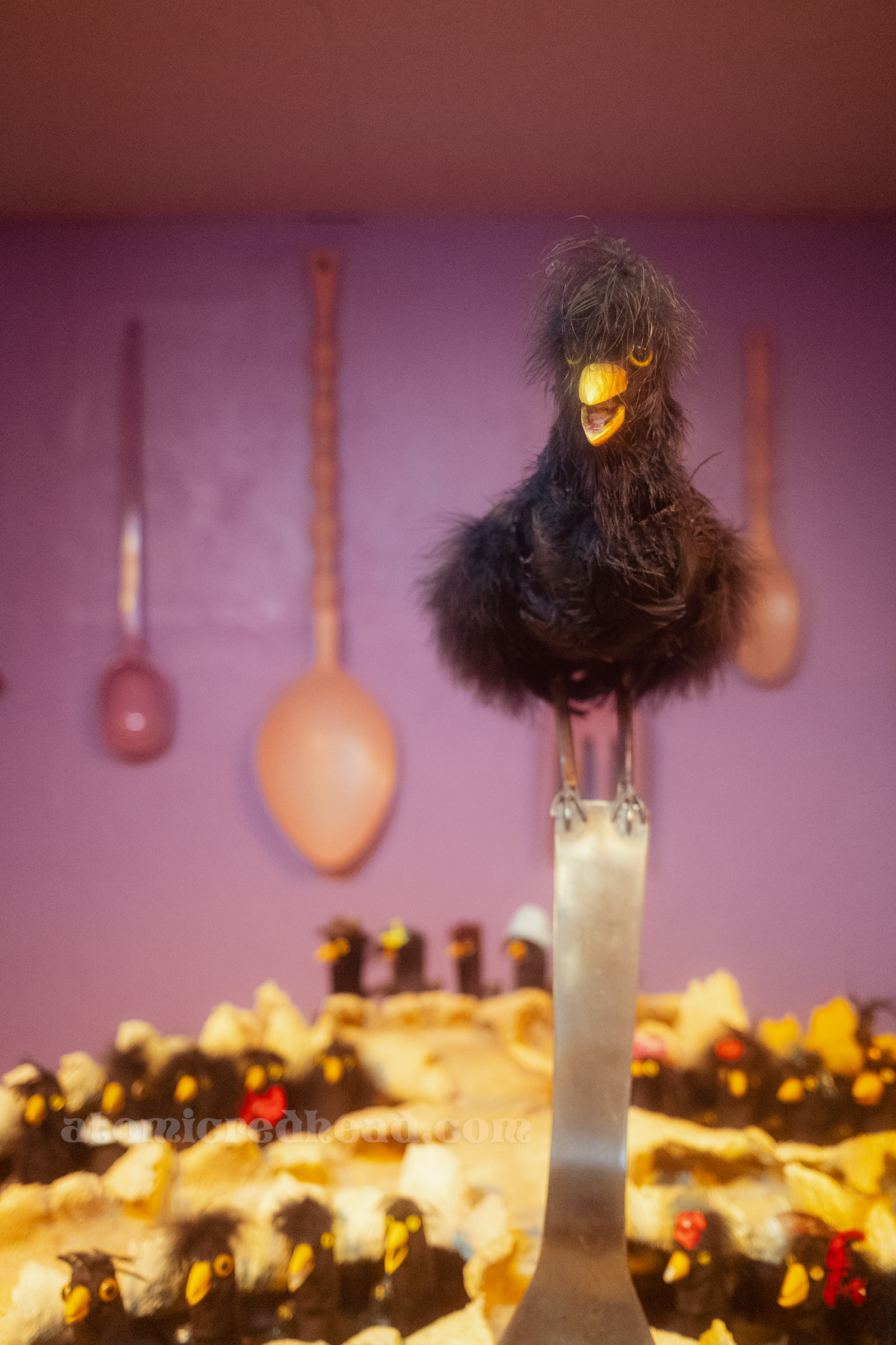 A large black bird sits on a large fork that is stuck in a massive pie with many smaller black birds popping out of.