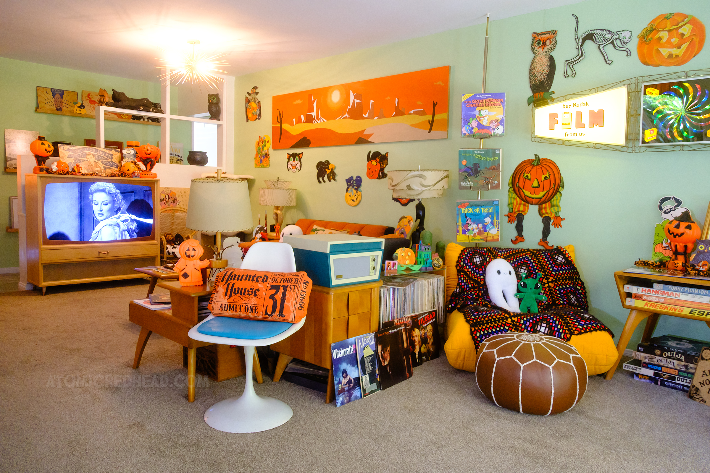 Overall of our living room. In the foreground a console holds a record player and records, next to it is a yellow chair, in the background is the view of our TV with House on Haunted Hill on the screen. Hanging on the wall are various diecut Halloween decorations.