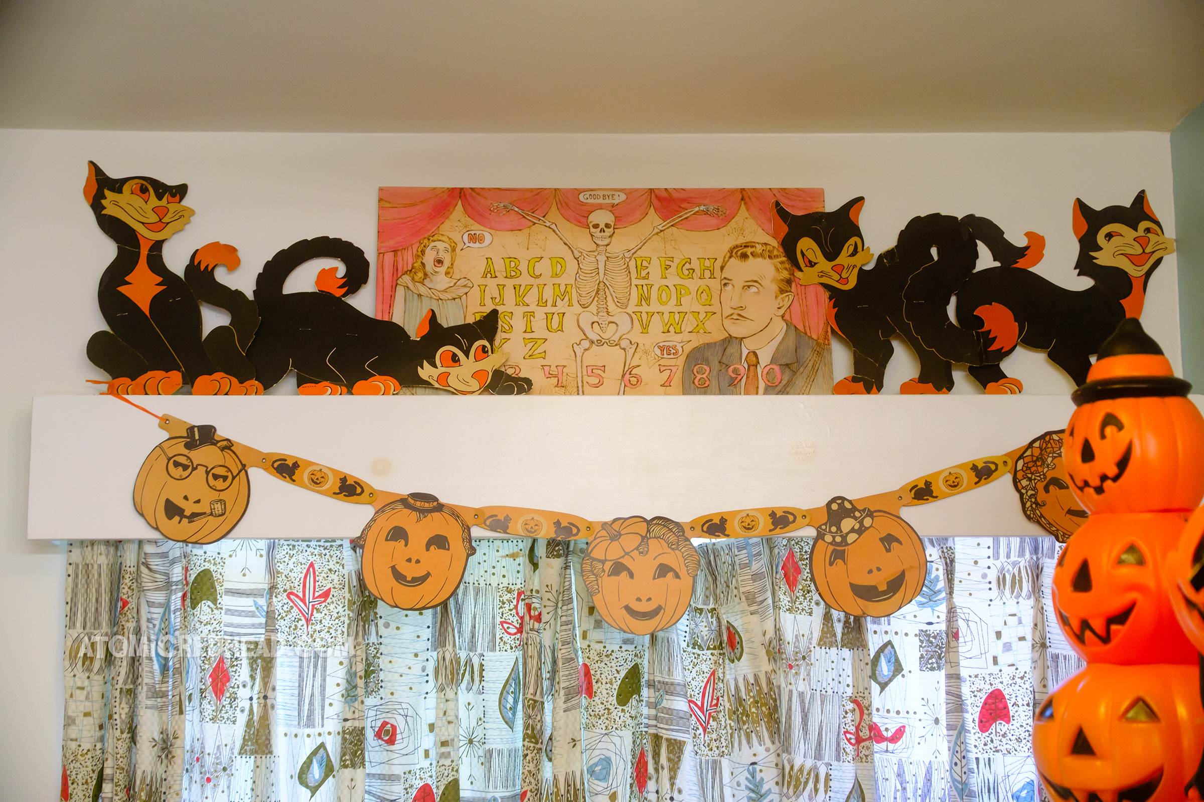 A spirit board themed to House on Haunted Hill sits on the valance above a window, also by it are four die cut black cats, and below a jack o'lantern banner hangs.