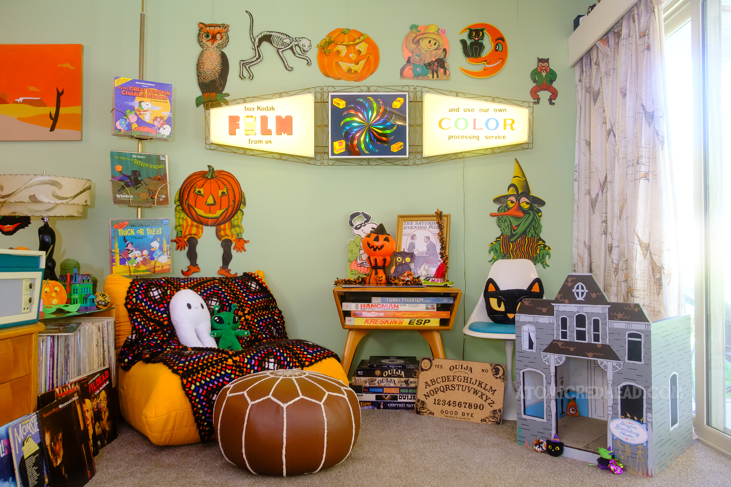 Overall of a part of our living room, hanging on the wall are multiple die cut Halloween decorations, including an owl, a skeleton cat, jack o'lantern, and a cat with a moon. Below are various Ouija games stacked on top of each other.