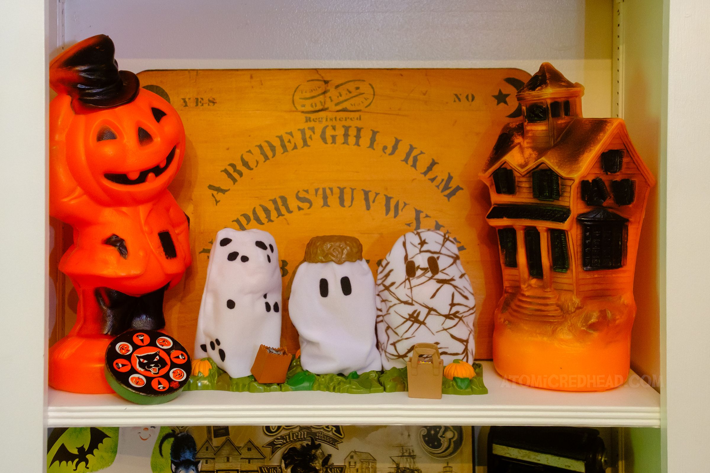 View of one of the shelves, on the left is a blow mold scarecrow, in the middle are Peanuts action figures of the kids dressed as ghosts, on the right is a blow mold haunted house and behind all of it is an antique Ouija board.