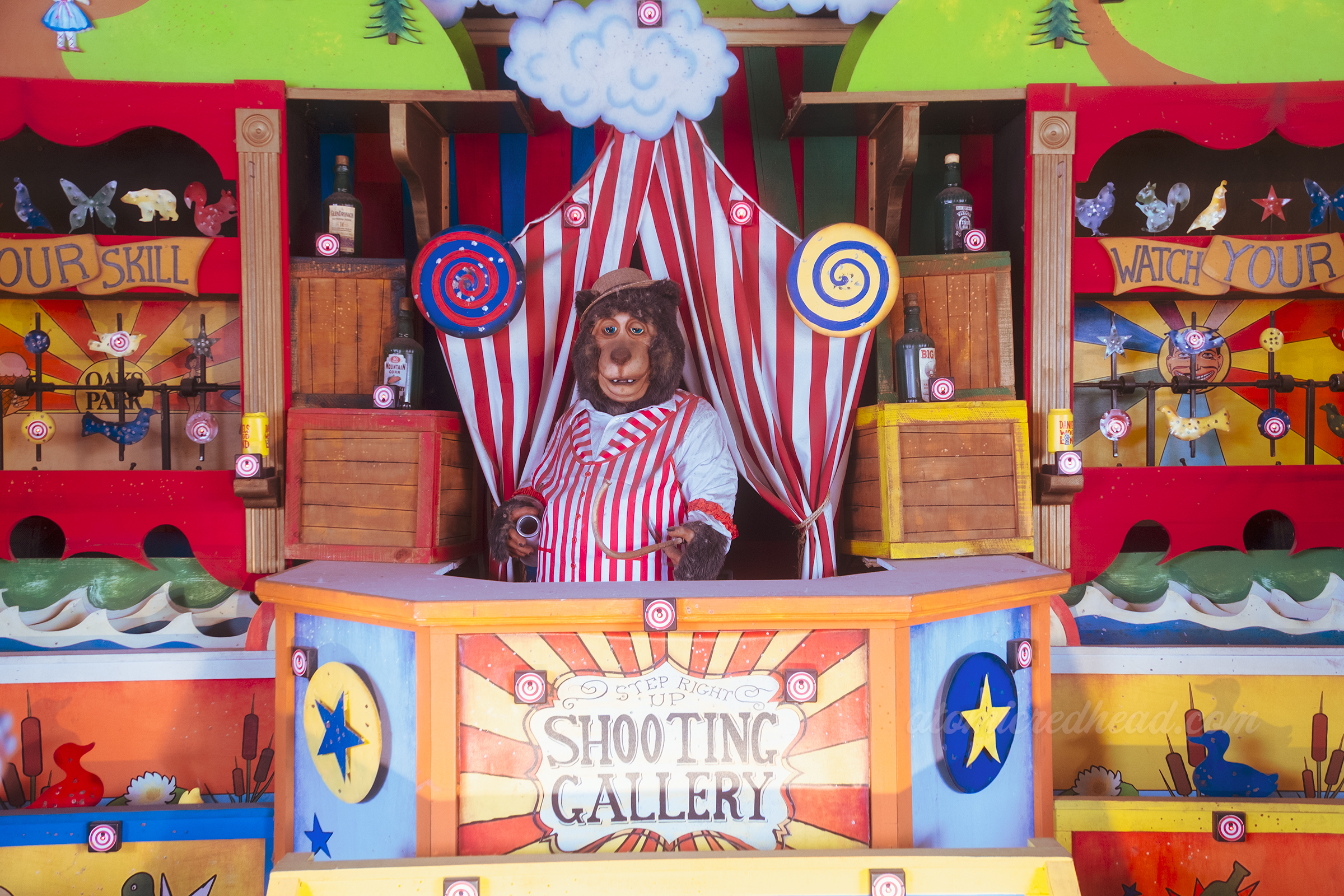 An animatronic bear dressed as a carnival barker stands in the middle of a shooting gallery.