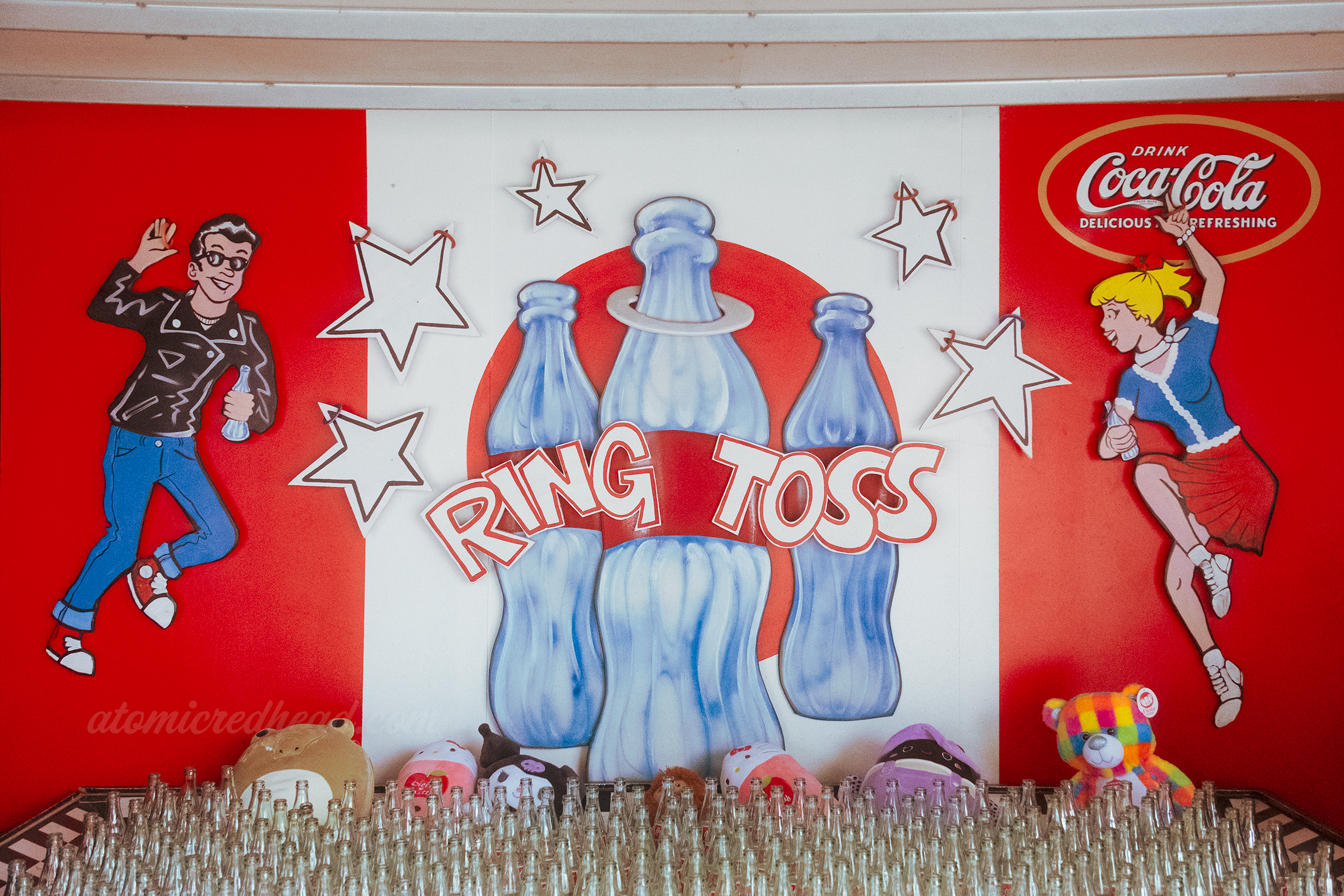 The ring toss game, with a collection of bottles at the bottom, above a hand painted mural features a 50s style couple, of a man in a black leather jacket and a girl in a skirt dancing.