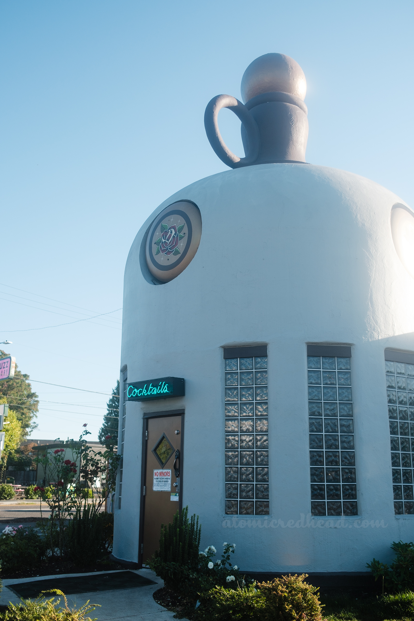Overall of the Sandy Jug, which is a building shaped like a jug, painted a pale cream. The very top is painted brown, with a gold stopper. Around the shoulder of the bottle are gold circles with roses painted in the middle. A door sits in the middle, with a blue neon sign reading "Cocktails" above, on either side of the door are windows of glass blocks.