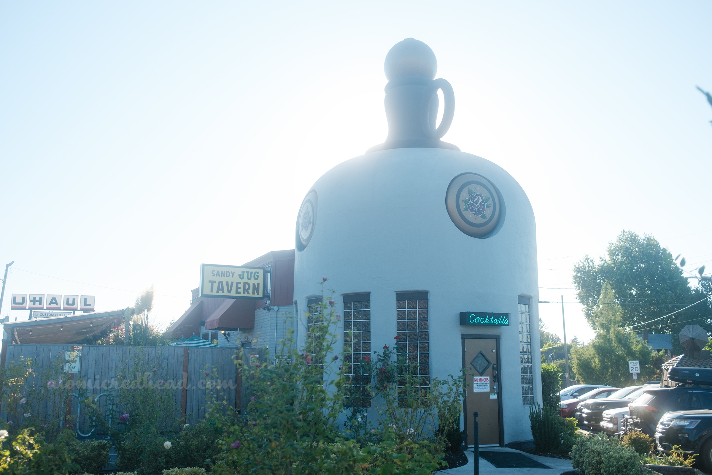 Overall of the Sandy Jug, which is a building shaped like a jug, painted a pale cream. The very top is painted brown, with a gold stopper. Around the shoulder of the bottle are gold circles with roses painted in the middle. A door sits in the middle, with a blue neon sign reading "Cocktails" above, on either side of the door are windows of glass blocks.