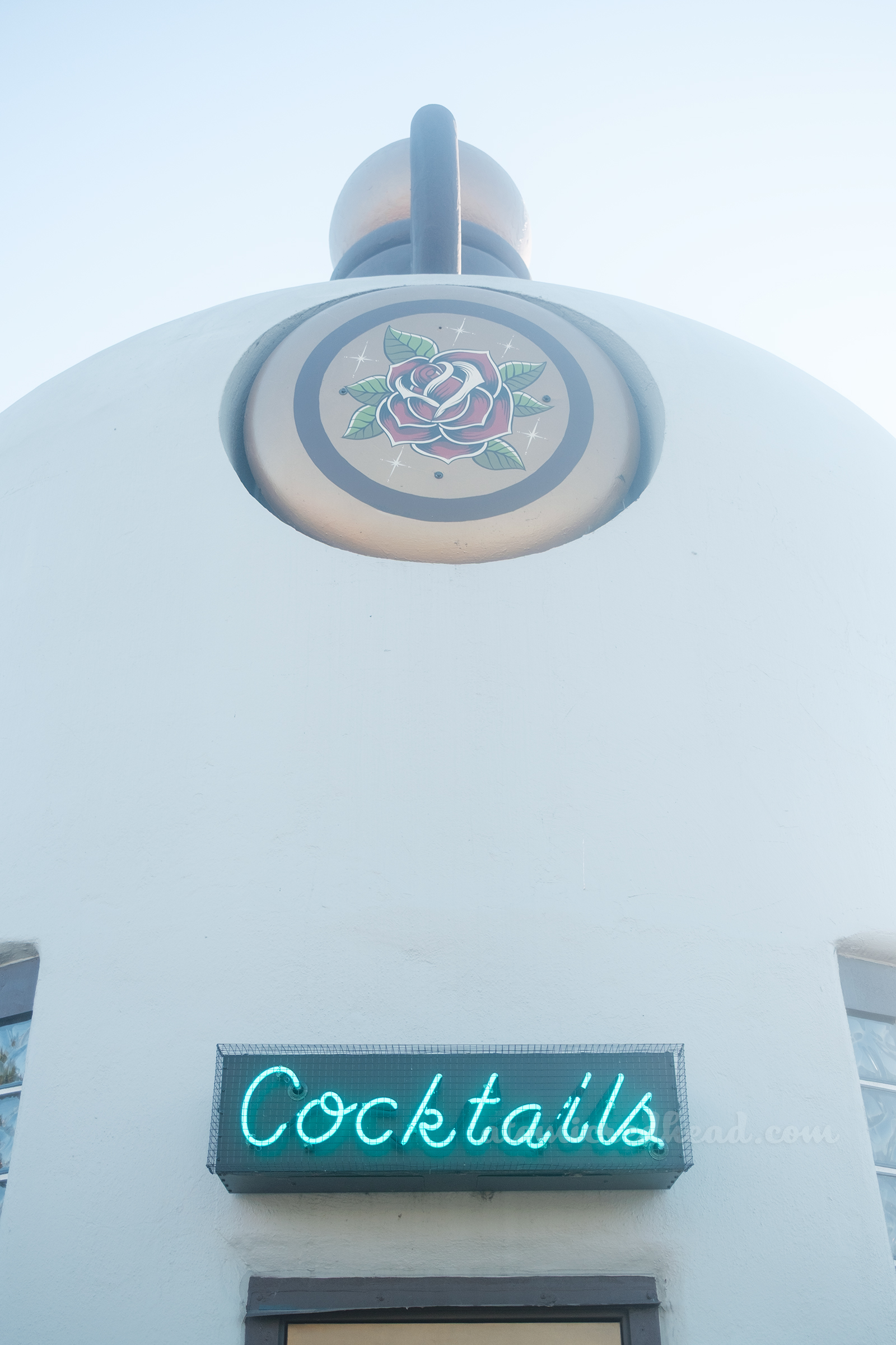 Overall of the Sandy Jug, which is a building shaped like a jug, painted a pale cream. The very top is painted brown, with a gold stopper. Around the shoulder of the bottle are gold circles with roses painted in the middle. A door sits in the middle, with a blue neon sign reading "Cocktails" above, on either side of the door are windows of glass blocks.