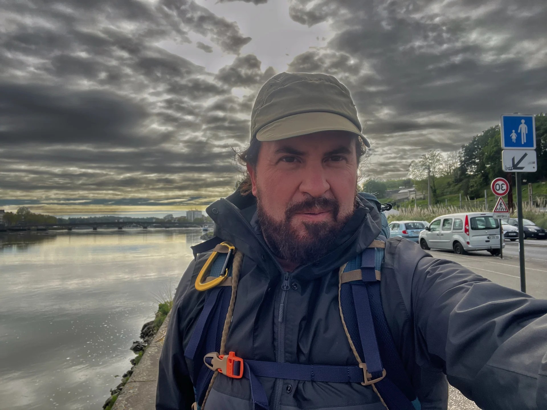Pilgrim wearing a backpack beside a river under a dramatic cloudy sky on the Camino de Santiago