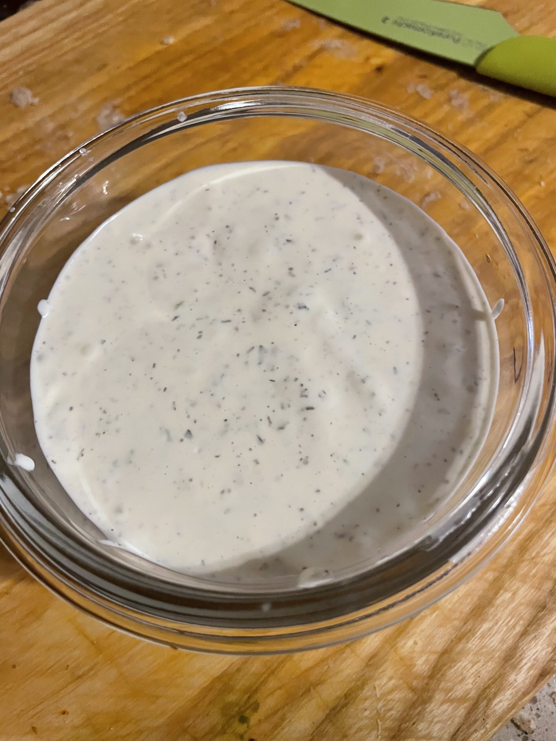 a glass bowl of white dip speckled with herbs on a cutting board with a green paring knife in the background