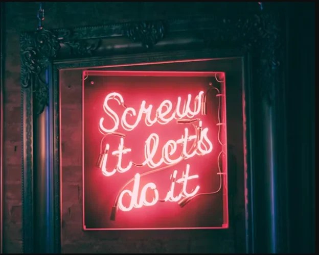 Neon sign with pink lighting that says "Screw it let's do it" displayed in a dark vintage frame on a brick wall