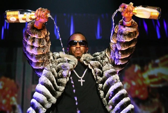 Diddy wearing an oversized fur coat with arms raised on stage, serving iconic fashion and hip-hop mogul energy.