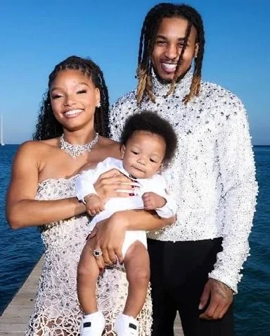 Halle Bailey smiling with DDG and their baby Halo in a family portrait by the water.