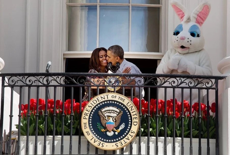 Barack and Michelle Obama waving from the White House balcony with a person in an Easter Bunny costume