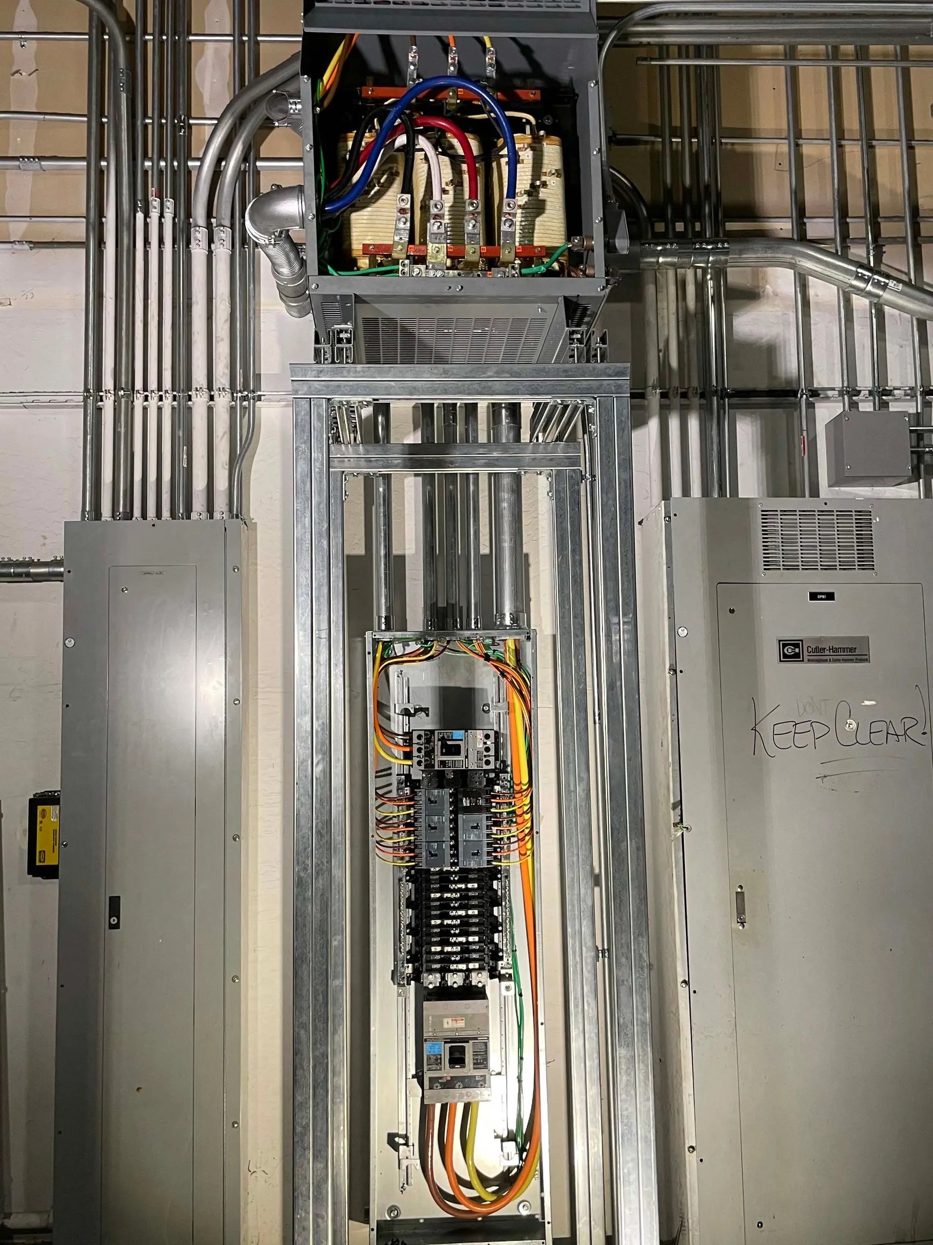An open electrical panel with exposed wiring and circuit breakers is mounted on a wall, surrounded by conduit pipes and closed metal cabinets. Bright lighting reveals detailed components—typical of work performed by skilled electricians San Jose trusts.