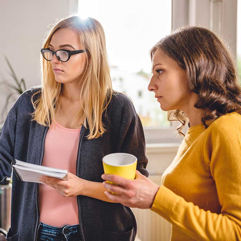 Two colleagues discussing work, one holding documents and the other a cup, in a brightly lit office setting.