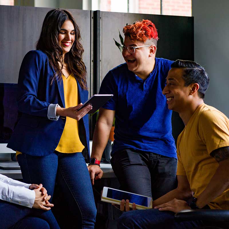 Three coworkers sharing a light-hearted moment, with one holding a tablet and two others laughing and enjoying the conversation in a casual office setting.