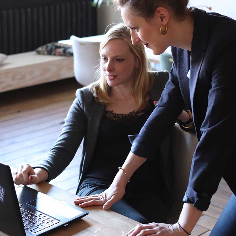 Two professional women collaborating at a laptop in a casual office setting.