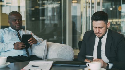 Two businessmen in suits sit on a couch with coffee cups, one using a tablet and the other looking at his phone. A notebook and pen are on the table in front of them in a modern office setting designed with network security in mind. This workspace features business network segregation and dedicated Guest Wi-Fi to ensure data protection for all users.