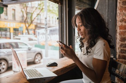 A woman sits by a window in a café, looking at her smartphone, with an open laptop and computer mouse on the wooden counter in front of her—illustrating the growing trend of using personal devices for work as part of modern BYOD practices.