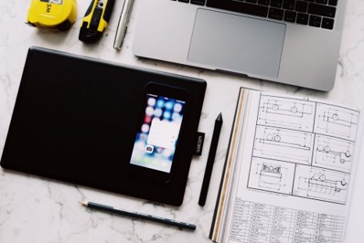 A workspace with a laptop, drawing tablet, smartphone, pen, pencil, measuring tape, utility knife, and technical drawings on a marble surface—ideal for professionals who prioritize network security while working on complex projects or managing their business network.