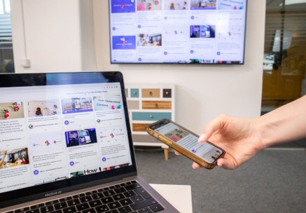 A person holds a smartphone in front of a laptop, both displaying similar content—an example of employees using personal devices for work. In the background, a large screen on the wall also shows related digital content. The setting appears to be an office or workspace.