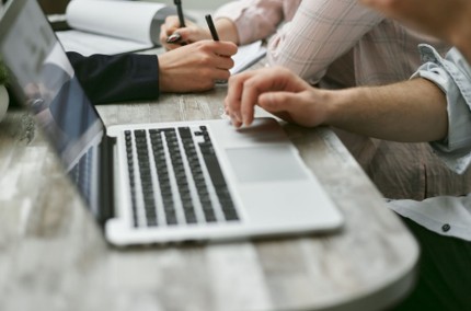 Two people sit at a wooden table working together; one types on a laptop while the other writes in a notebook, demonstrating careful employee computer security practices. Papers and a pen are visible on the desk.
