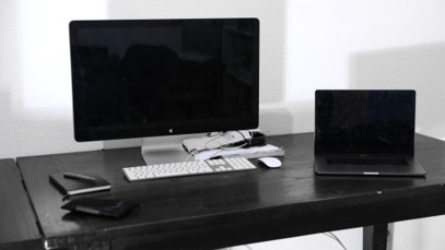 A black desk with a large monitor, keyboard, mouse, closed laptop, and some wallets or small cases in a minimalist office setting—an ideal workspace for those looking to maximize their business computers’ efficiency and extend computer lifespan.