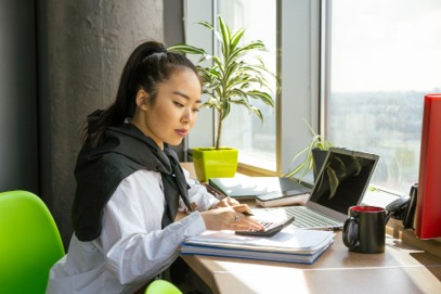 A woman sits at a desk with a laptop, calculator, and papers, focused on IT budget planning for her company. She is working diligently—with a plant and coffee mug nearby—next to a window with daylight streaming in, making the most of small business technology to optimize her Small Business IT Budget.