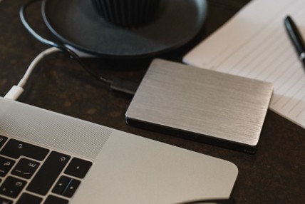A close-up of a laptop connected to an external hard drive, with a lined notepad and a pen nearby on a dark surface, suggests careful consideration of data deletion and managing cloud data retention.