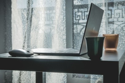 A laptop, computer mouse, coffee cup, and candle sit on a table by a window with sheer curtains, letting in soft natural light—a serene setting that might inspire you to consider how long business computers last as you work.