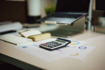 A calculator rests on printed charts beside a notebook, keyboard, and a laptop on a white desk in an office setting, reflecting the meticulous approach needed for Small Business IT budget planning.
