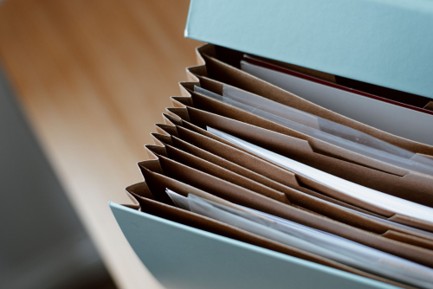 Close-up of an open accordion file organizer with several paper documents sorted in brown tabbed sections, representing organized records for cloud service data. The background shows a blurred wooden surface.