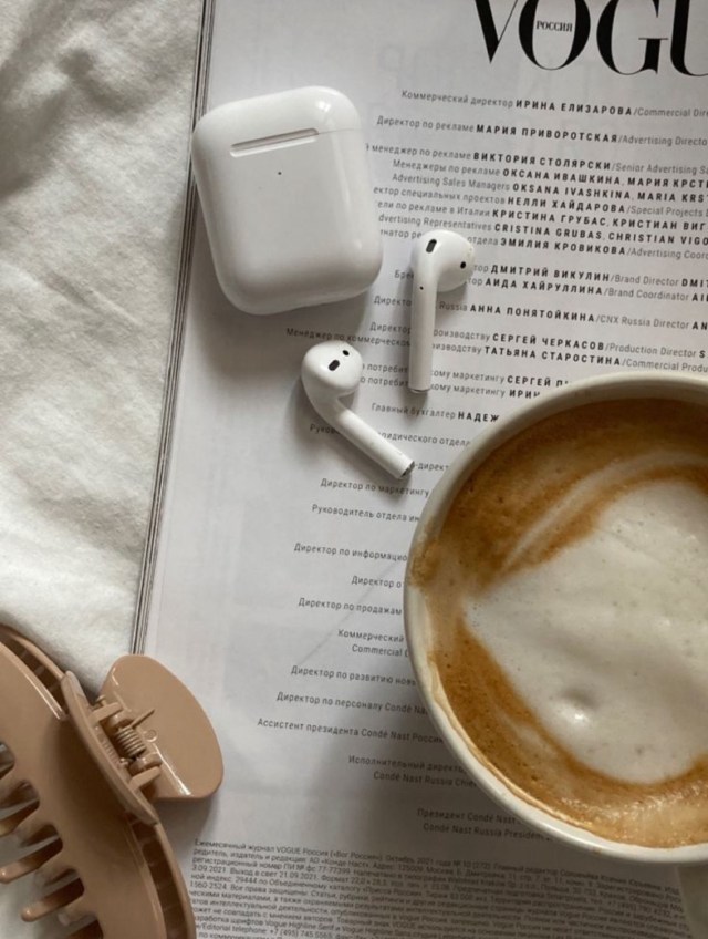 Flat lay of a coffee cup with frothy milk, wireless earbuds, and an AirPods charging case on top of a magazine page.