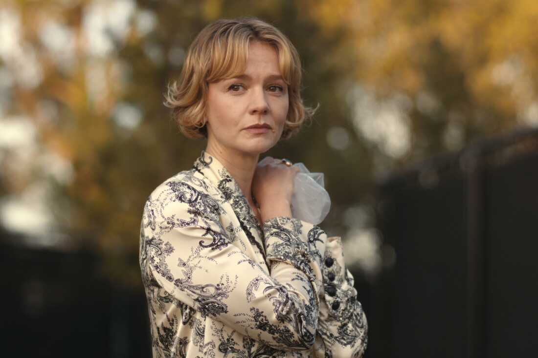 "Woman with short blond hair wearing a patterned jacket stands outdoors in front of a blurred background of autumn trees, holding a face mask in her hand and looking thoughtfully into the distance."