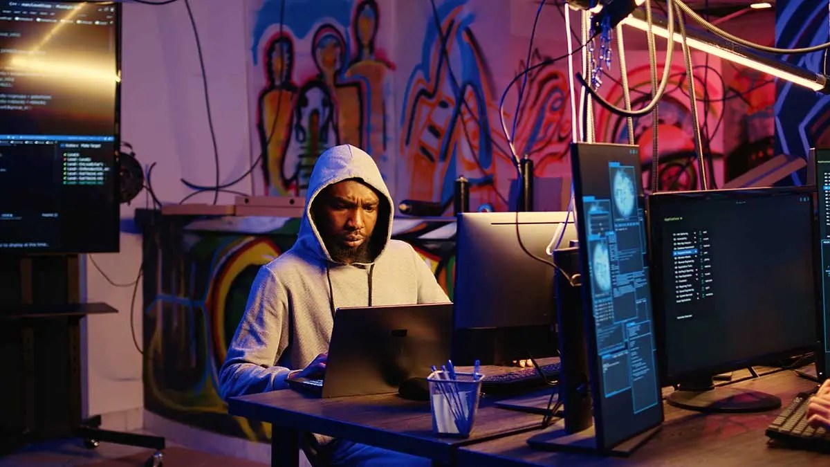 Cybersecurity hacker in a dark room working on multiple monitors with code and security data displayed on screens.