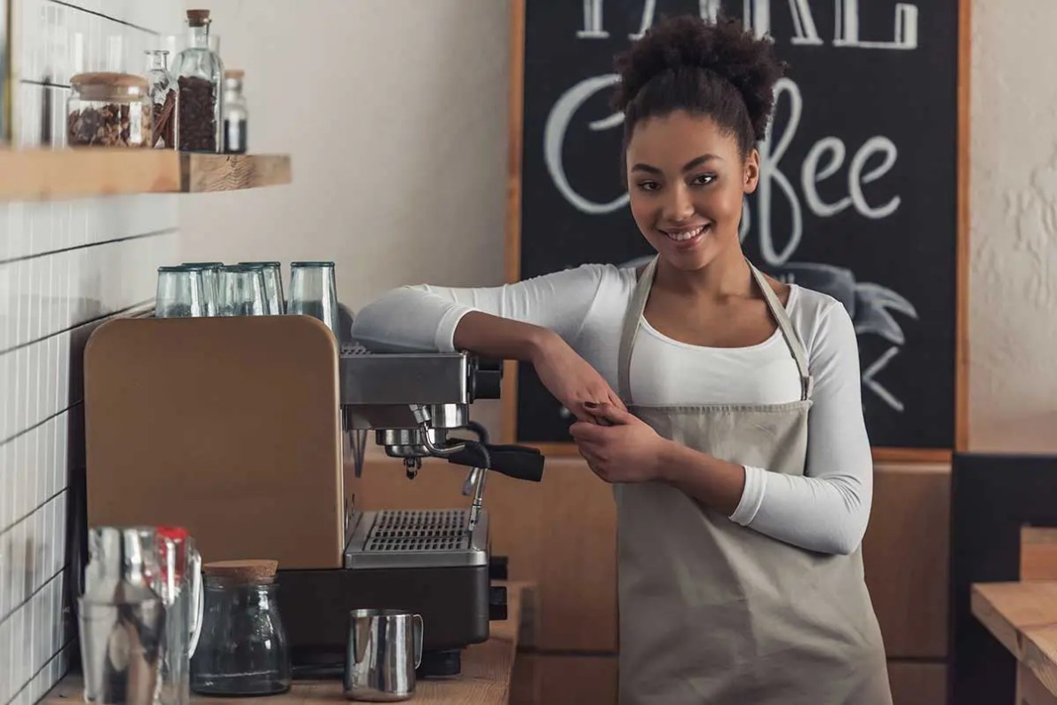 Friendly Barista – Elevate Customer Experience Through Human Connection A smiling barista in a beige apron standing confidently beside an espresso machine in a cozy coffee shop, ready to serve customers.