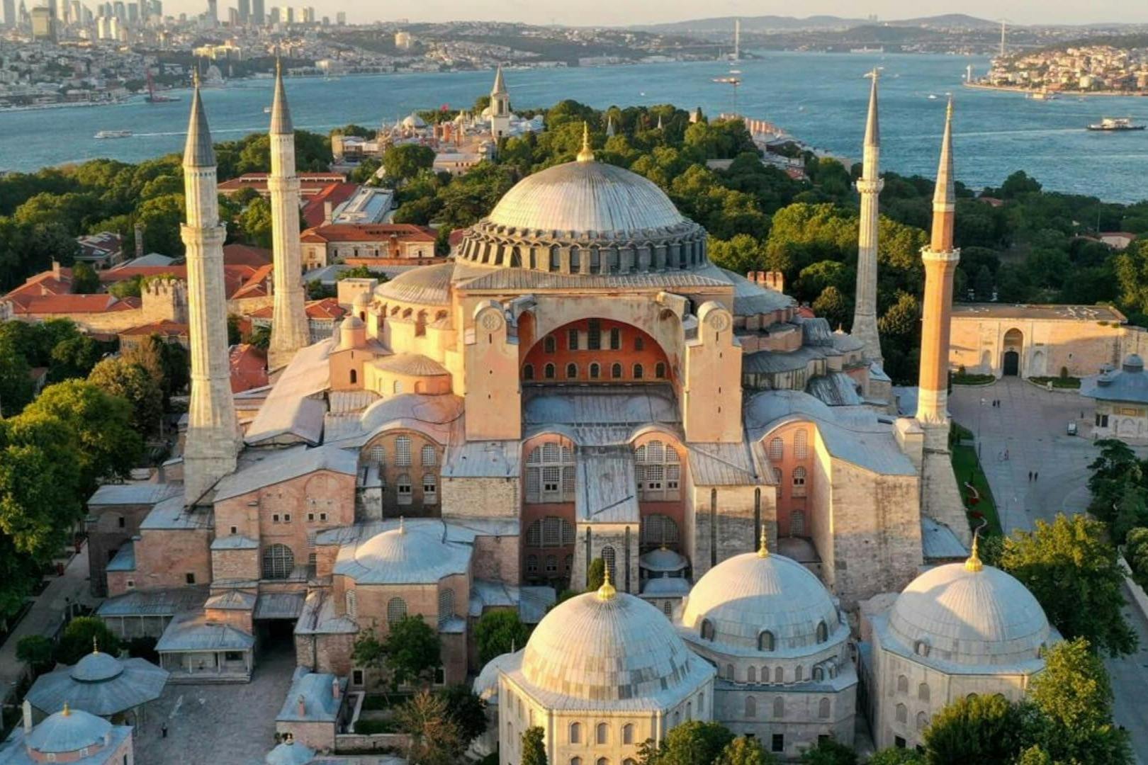 Aerial view of Hagia Sophia in Istanbul, featuring domes, minarets, and surrounding greenery, with a waterway in the background.