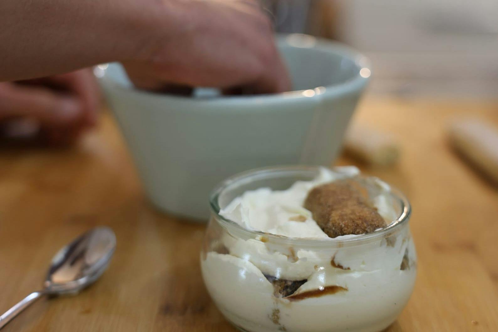 A hand dips into a large bowl while making tiramisu; a glass dish with layers of cream and ladyfinger biscuits is in the foreground.