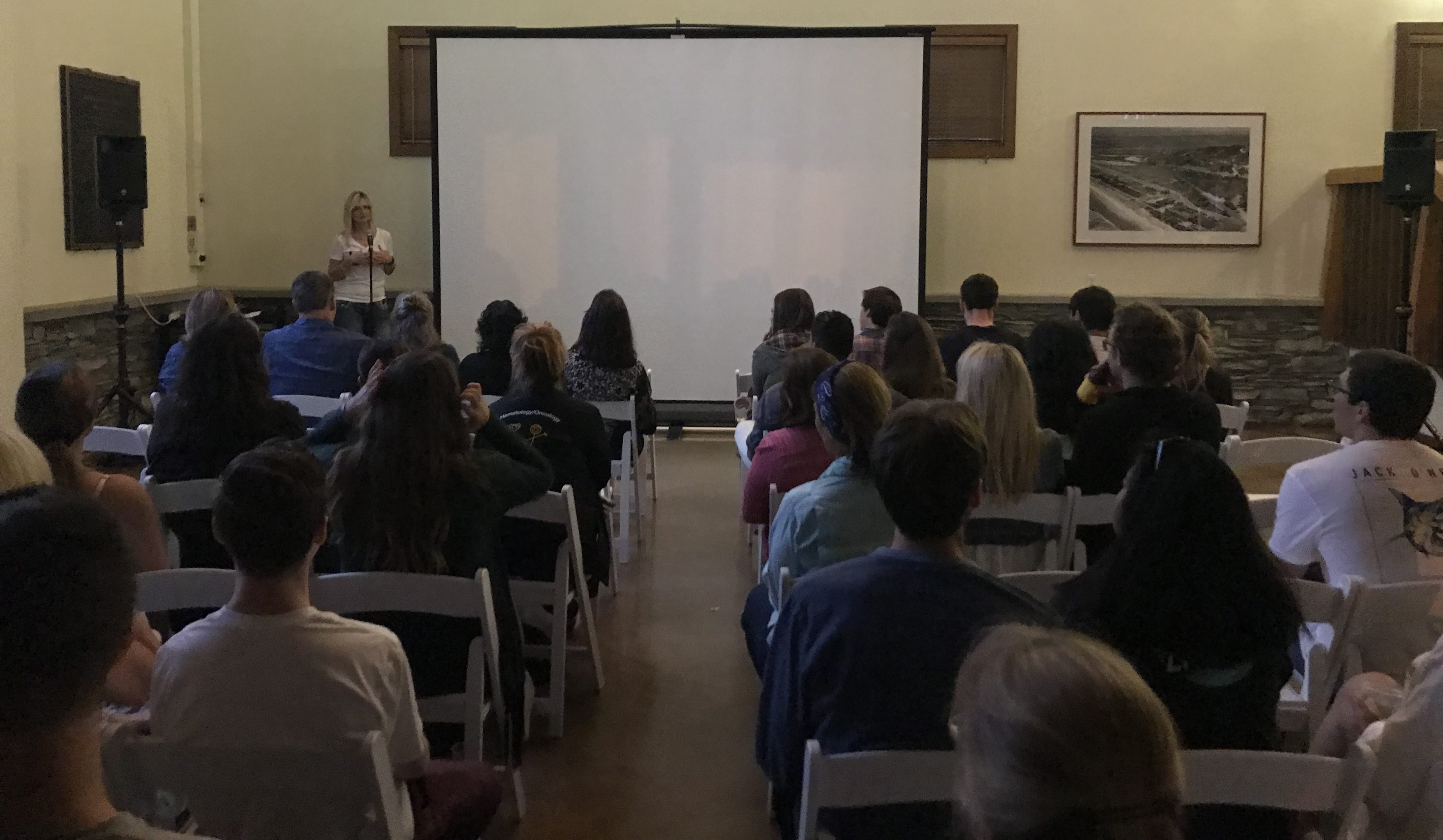 A woman speaks at the front of a dimly lit room to an audience seated on white chairs, facing a large blank projection screen. The setting suggests a discussion or support group about young adult cancer, with beige walls and framed pictures.
