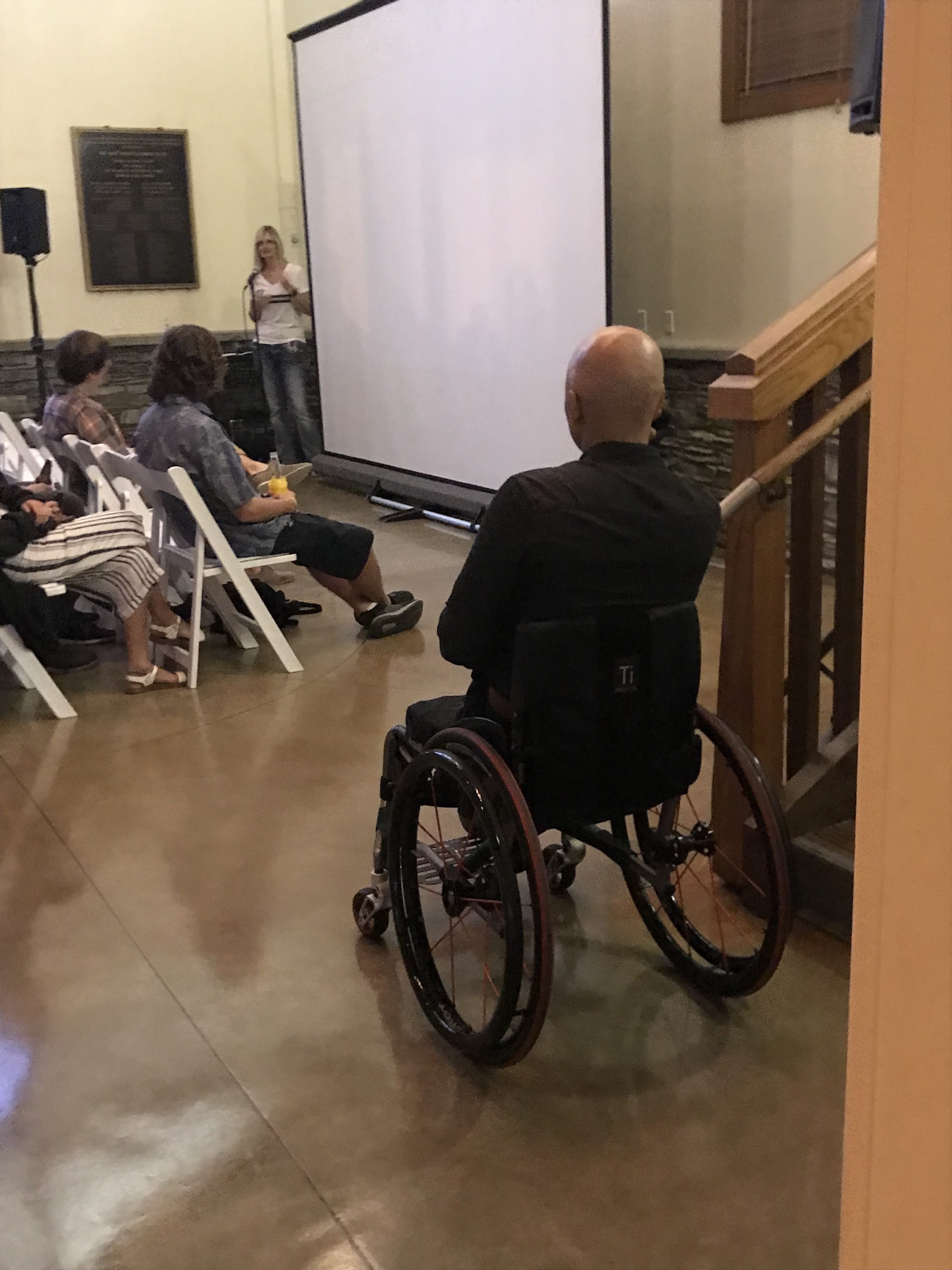 A man in a wheelchair faces a projection screen at an indoor event, with seated attendees and a woman speaking at a podium in the background.