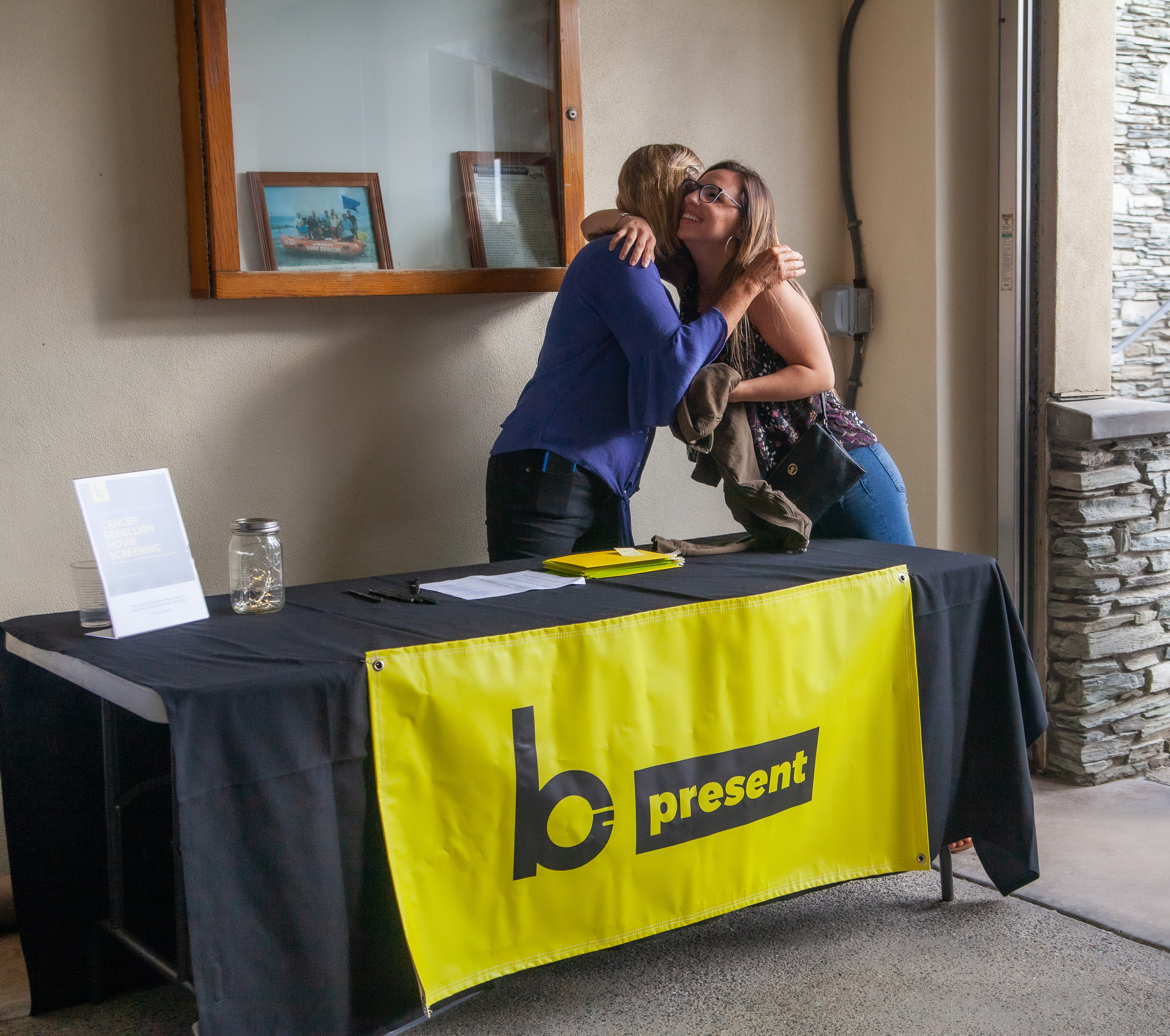 Two women greet each other with a hug at a check-in table covered by a bright yellow b-present banner in a lobby area, with a sign-in sheet and a donation jar on the table.